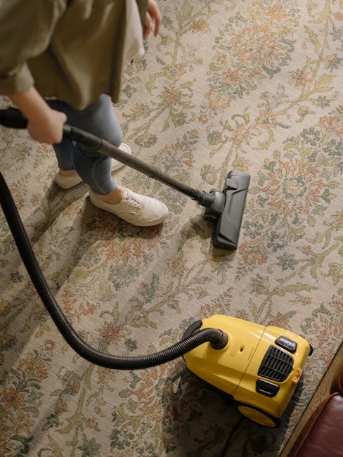 A person kneeling on a patterned carpet in a residential or commercial space is seen cleaning a yellow and black vacuum cleaner. The individual is using a cloth to wipe the inside of the vacuum's dust