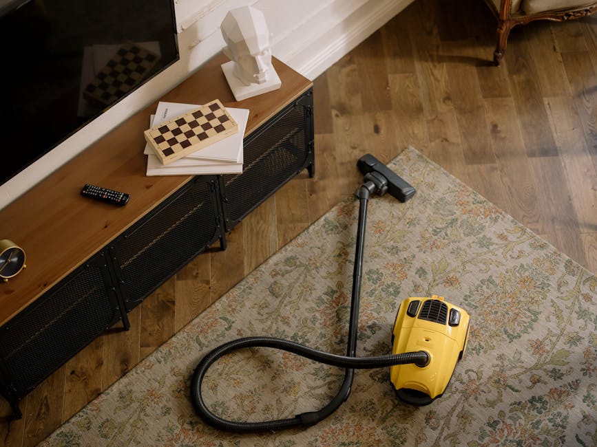 A woman using a yellow vacuum cleaner to deep clean a patterned beige and cream carpet in a living room. She is dressed casually in a beige jacket, jeans, and white sneakers. The room features wooden 