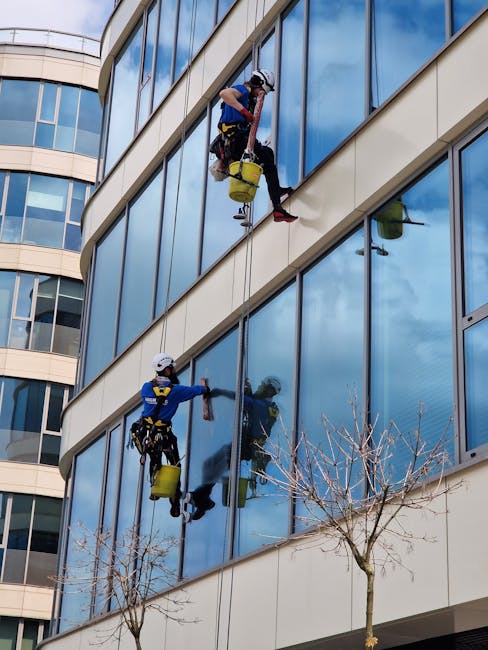 Two professional window cleaners are suspended on a scaffolding platform high above the ground, performing surface cleaning and detailing on the exterior glass facade of a modern multi-storey building